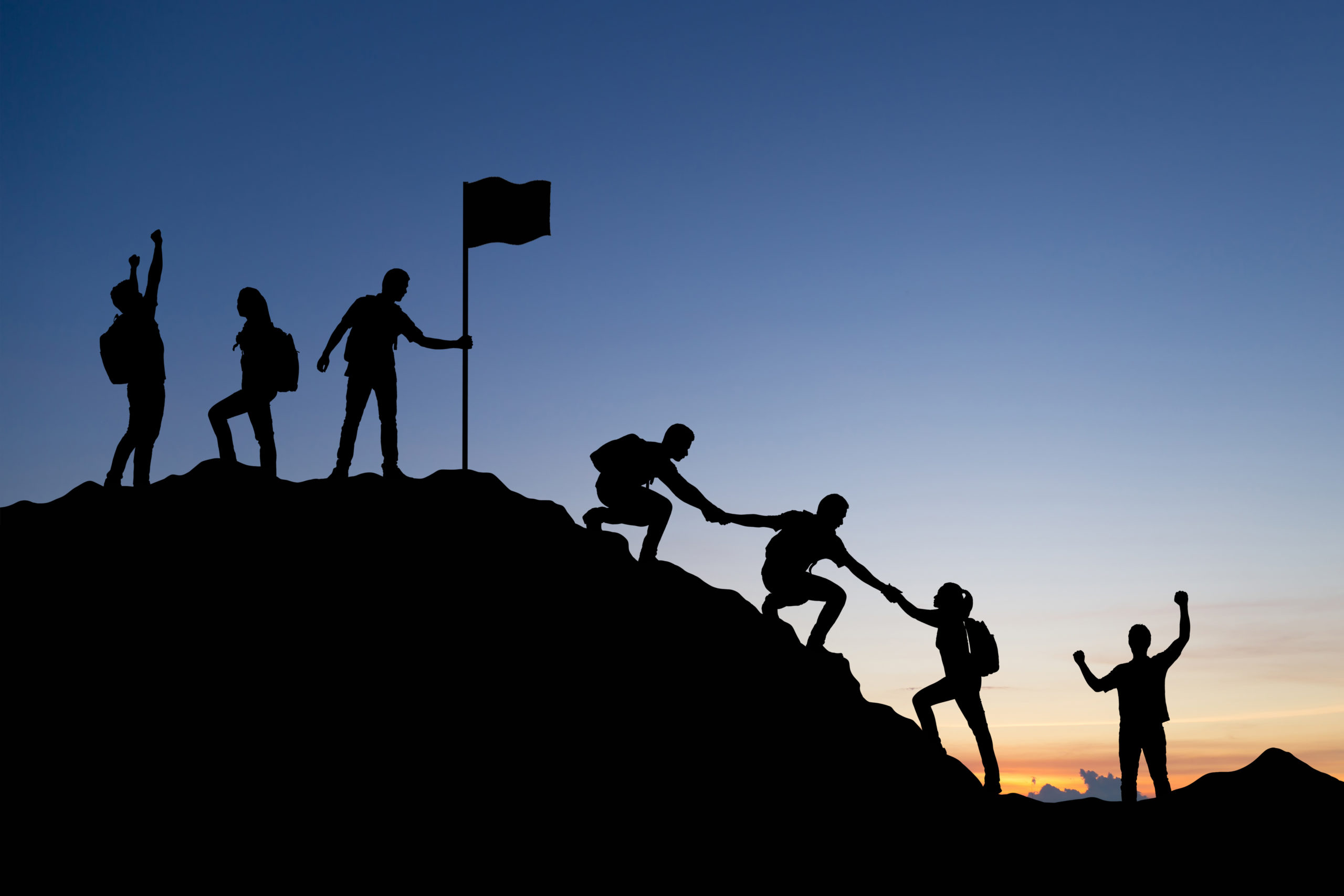 Silhouette of people helping each other hike up a mountain at sunset background. Teamwork, success and goal concept.